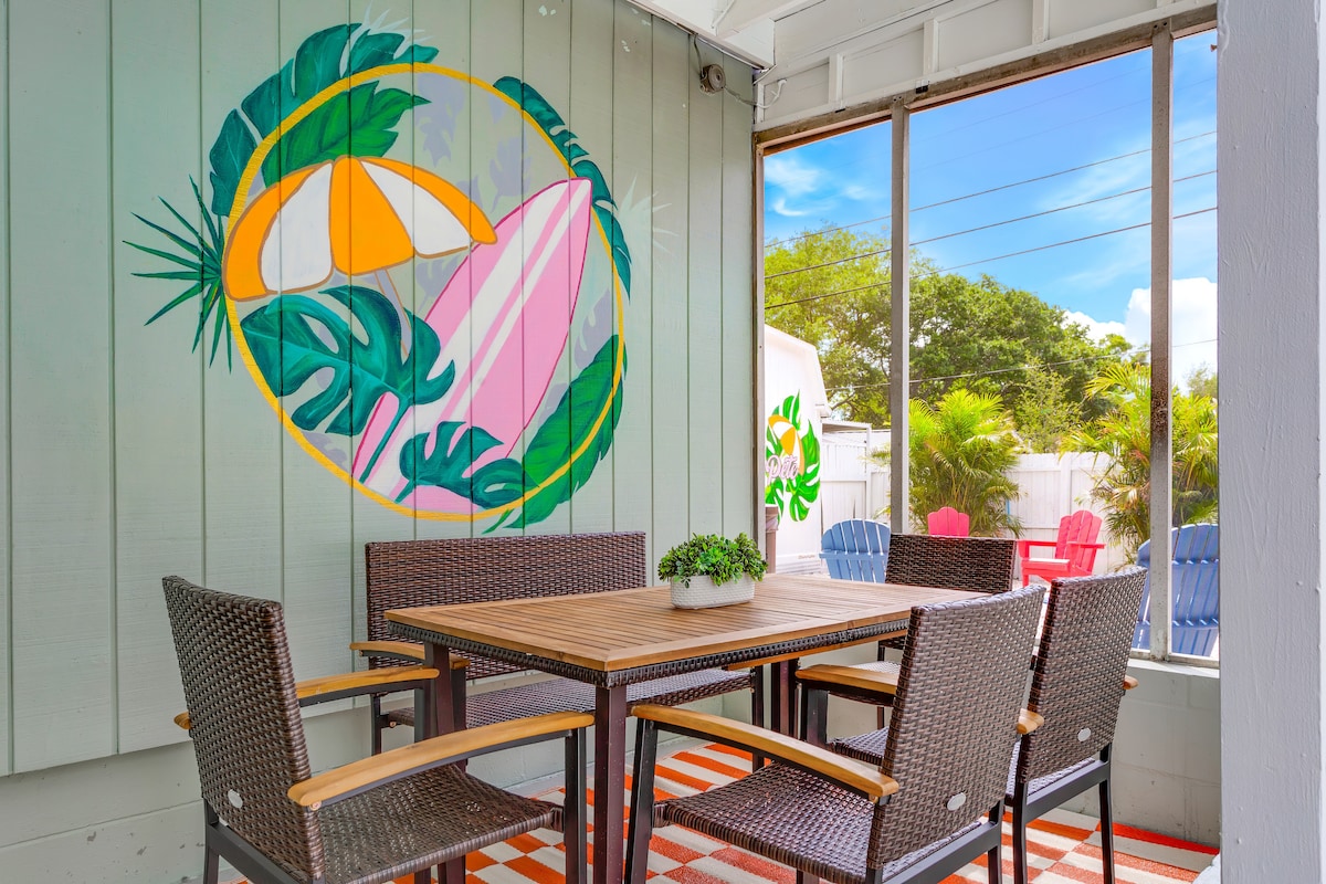 A sunlit sunroom features a wooden dining table surrounded by six woven chairs. A tropical-themed mural adorns the wall, depicting surfboards and palm leaves. Large windows and sliding glass doors offer views of the outdoor space, while a vibrant rug adds a pop of color to the floor.
