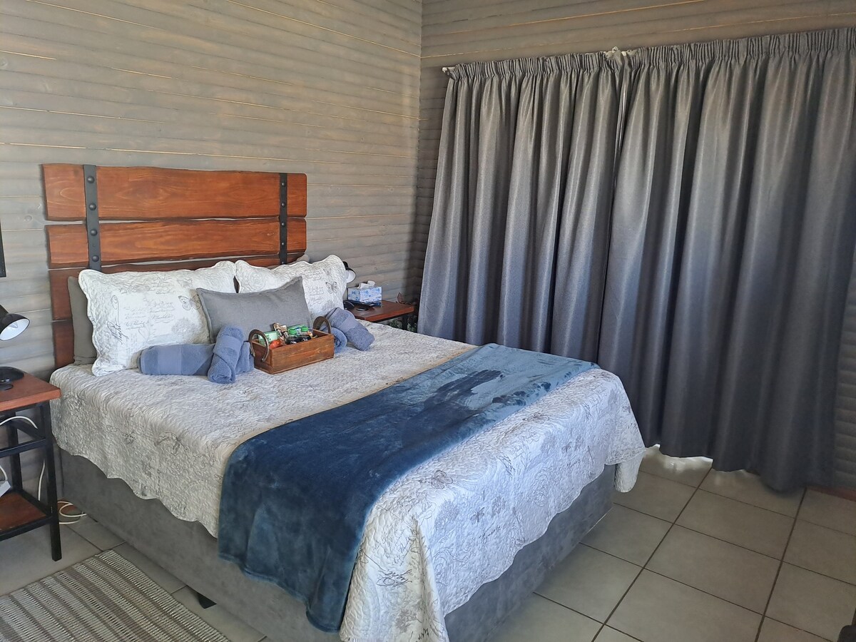 A spacious bedroom features a large bed adorned with a textured blanket and decorative pillows. Soft light filters through floor-length gray curtains, providing a calm ambiance. A wooden headboard adds a rustic element, while a bedside table displays a small basket and a lamp.
