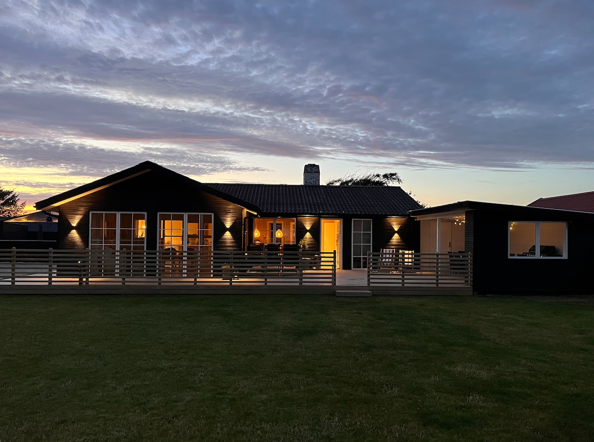 The exterior of the summer house is shown at dusk, with warm lighting emanating from multiple windows. A spacious wooden deck features a railing, and the home's architecture combines a traditional sloped roof with modern elements, set against a colorful sky.