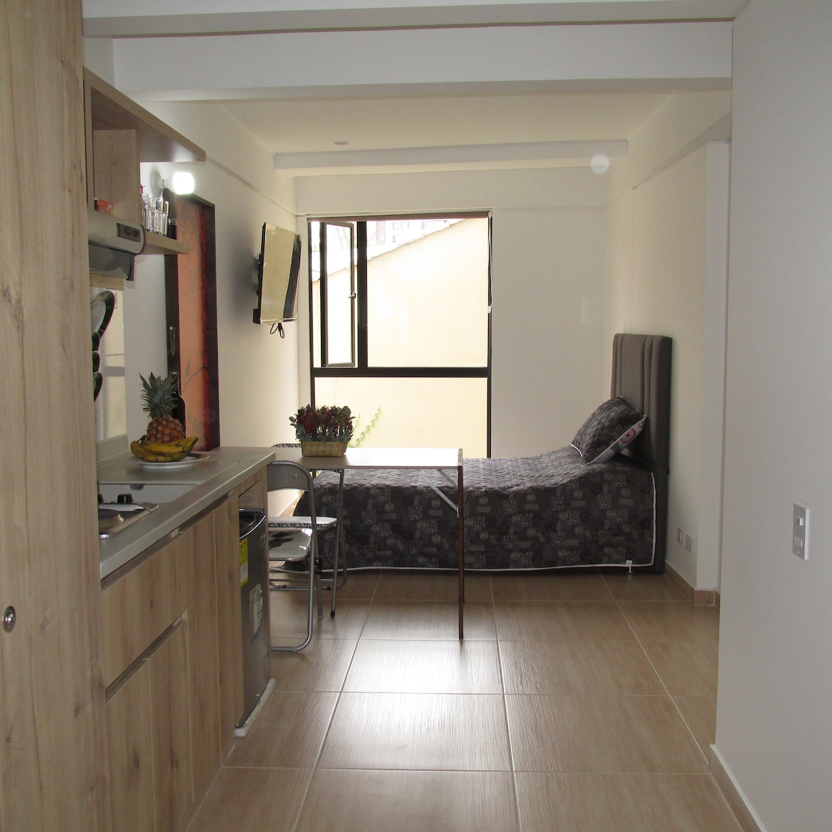 An open-plan layout features a compact kitchen area with modern appliances and a dining table. A single bed is positioned next to a large window, allowing natural light to fill the space. Neutral tones and simple decor contribute to a minimalist ambiance.