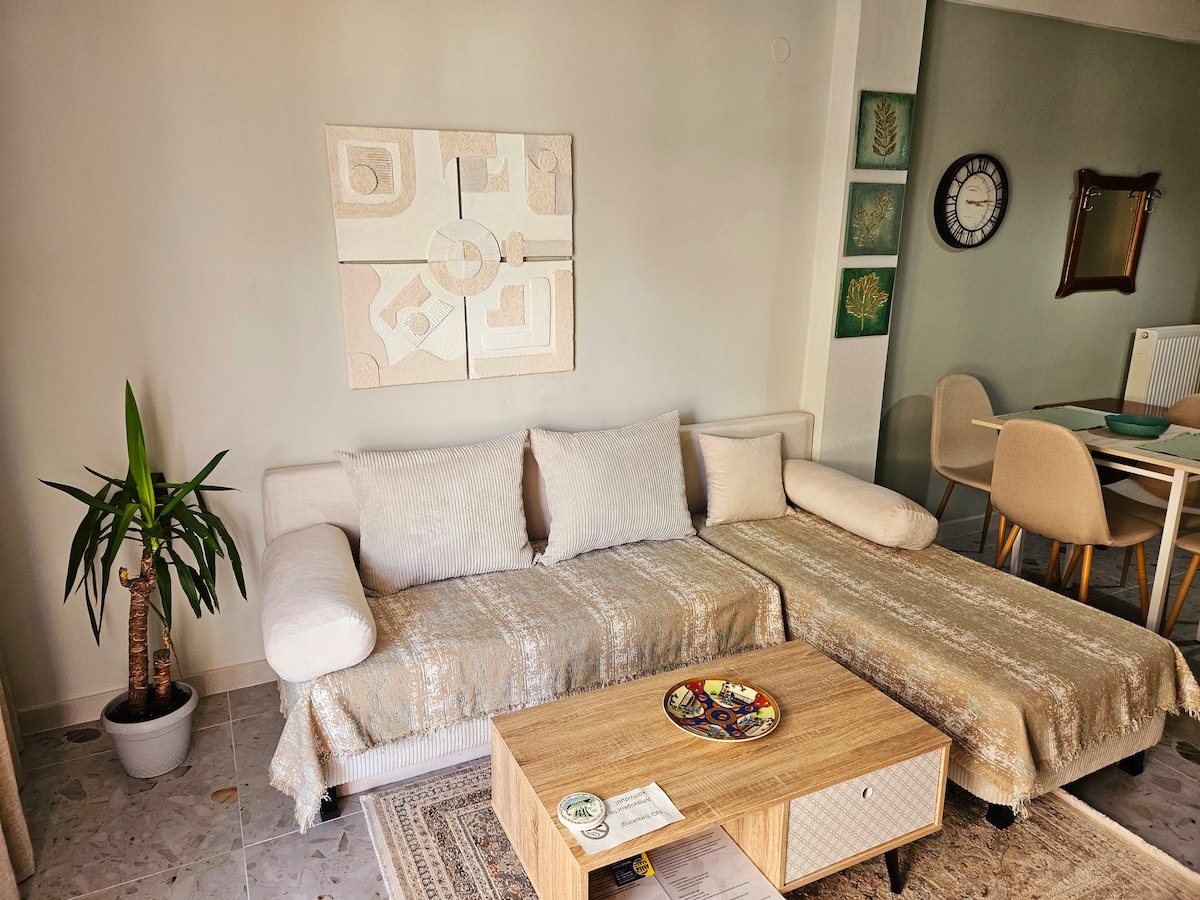 The living area is adorned with a light-colored sofa and a wooden coffee table, complemented by neutral textiles. A small plant adds a touch of greenery, while artwork and a circular wall clock enhance the decor. A dining table with chairs is visible in the background.