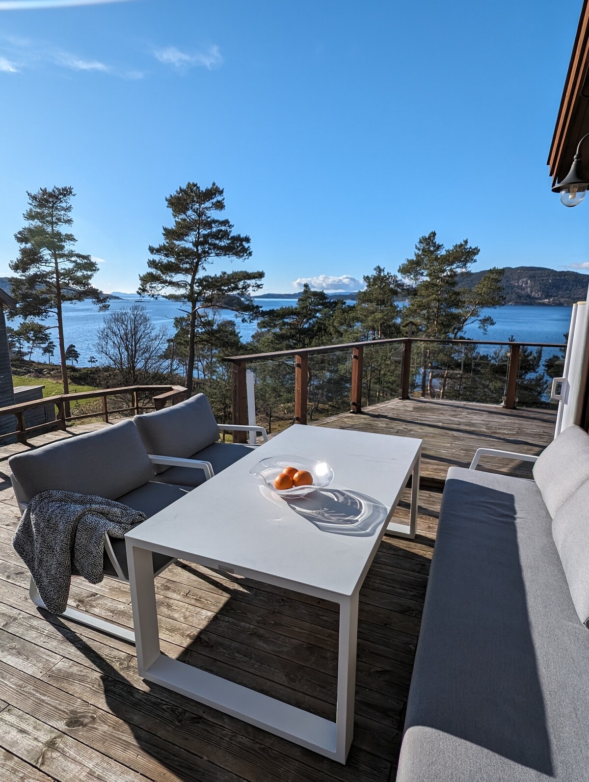 An outdoor terrace features a dining table and two armchairs set against a backdrop of clear blue skies and scenic waterfront views. A bowl of fruits sits at the center of the table, accompanied by a cozy throw draped over one chair.