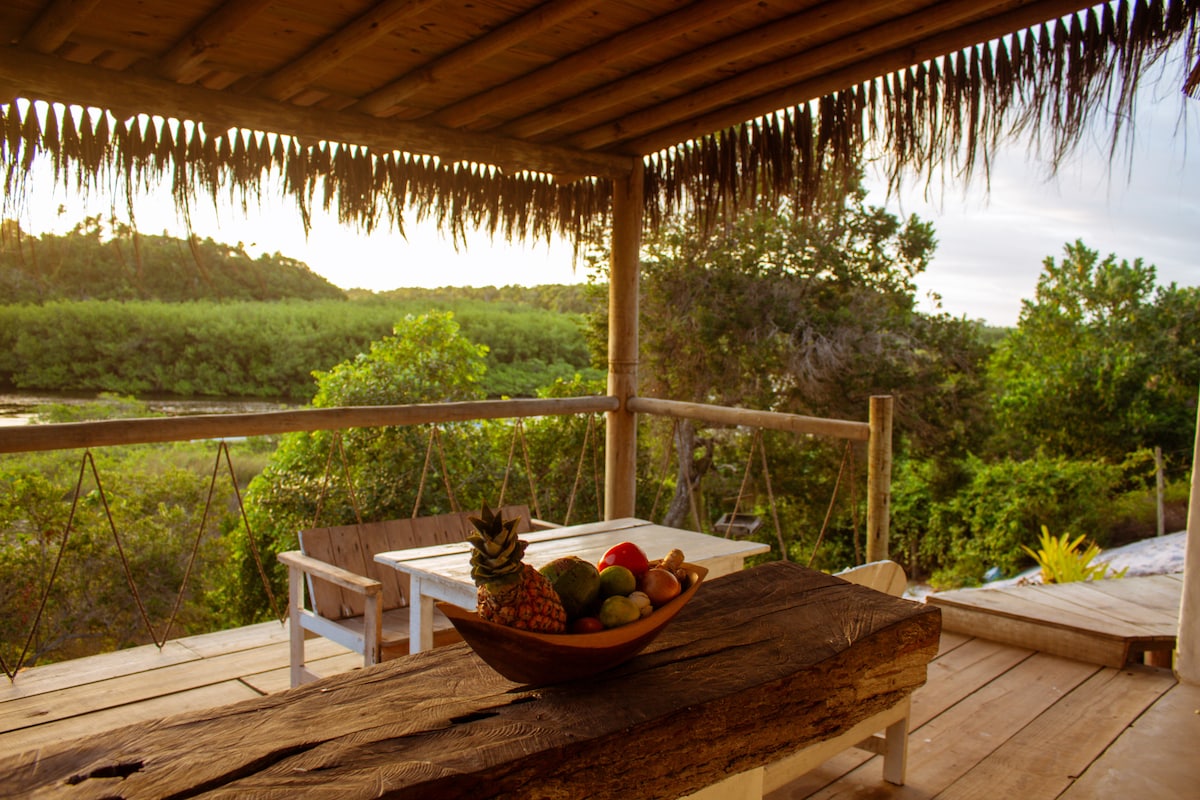 A wooden table adorned with a bowl of fresh fruits is positioned on a covered patio. Surrounding greenery and the tranquil river can be seen in the background, with palm fronds softly framing the space, creating a natural and relaxing environment.