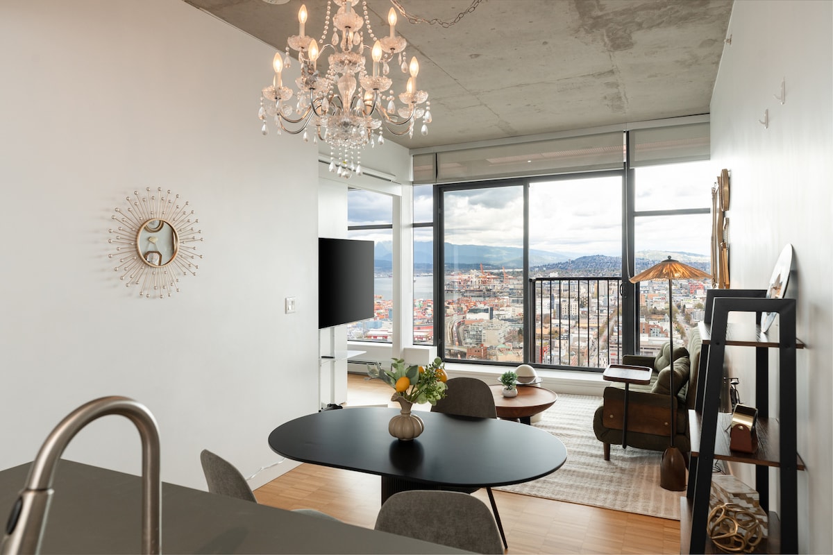 A stylish living area features large windows offering panoramic views of Vancouver. A round black dining table sits in the foreground, accompanied by a contemporary chandelier. A green armchair and wooden side table enhance the cozy ambiance, while a flat-screen TV is positioned nearby.