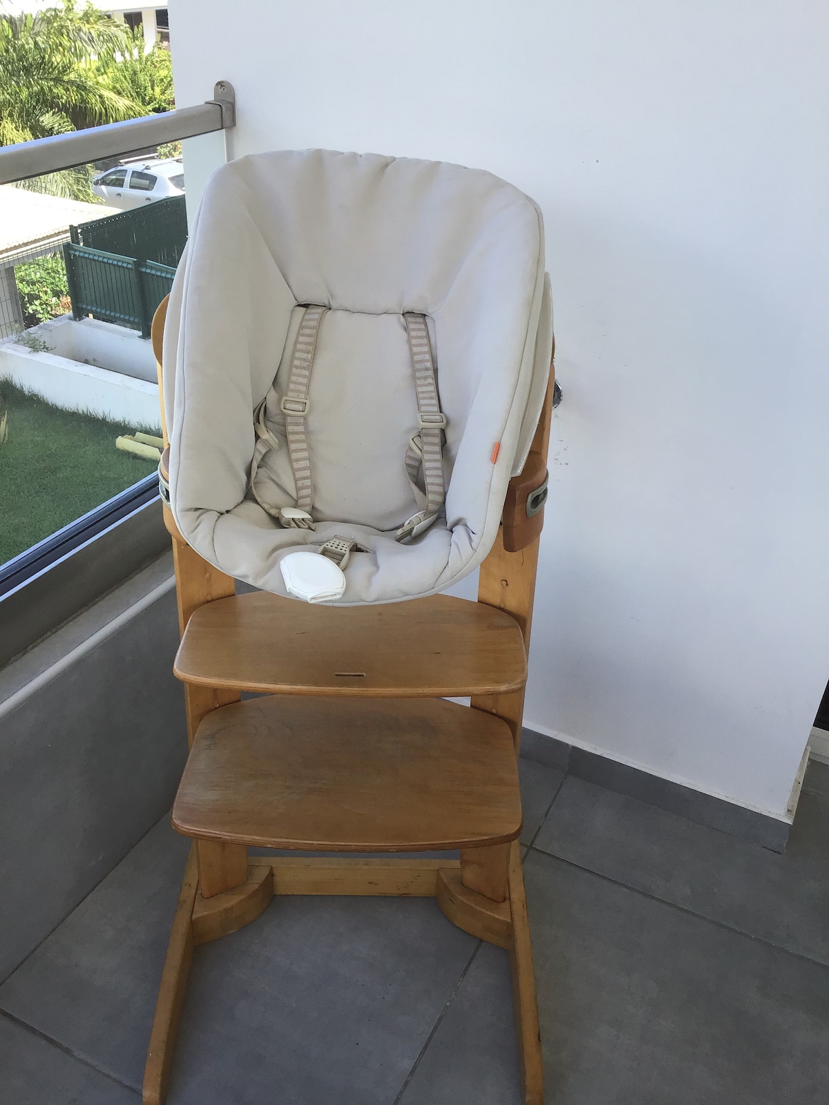 A wooden high chair is positioned on a balcony, showcasing a soft grey cushion. The chair is elevated with ample space below, and a backdrop of green grass and nearby structures can be seen through the glass railing.