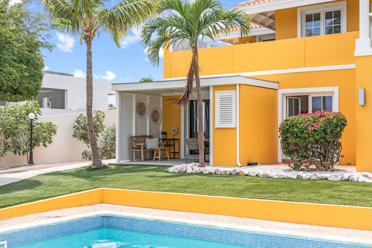 The image features a vibrant yellow villa with a spacious porch, surrounded by lush greenery and palm trees. A sparkling pool is situated in the foreground, complemented by neatly trimmed grass and colorful flowering plants, creating a serene outdoor atmosphere.