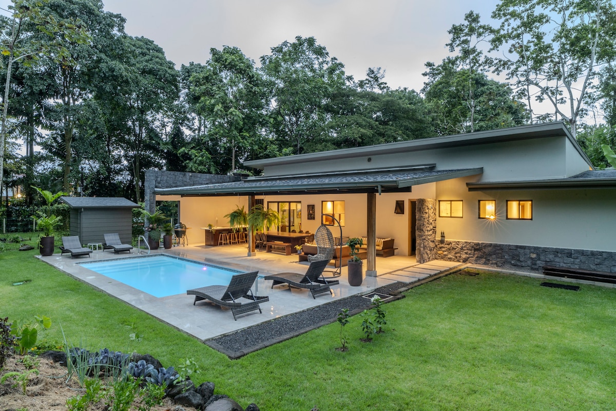 A modern home is set amidst lush greenery, featuring a spacious outdoor area with a crystal-clear pool. Comfortable lounge chairs are positioned around the pool, and large glass doors provide an inviting view into the interior. Tropical plants and manicured lawns enhance the serene environment.