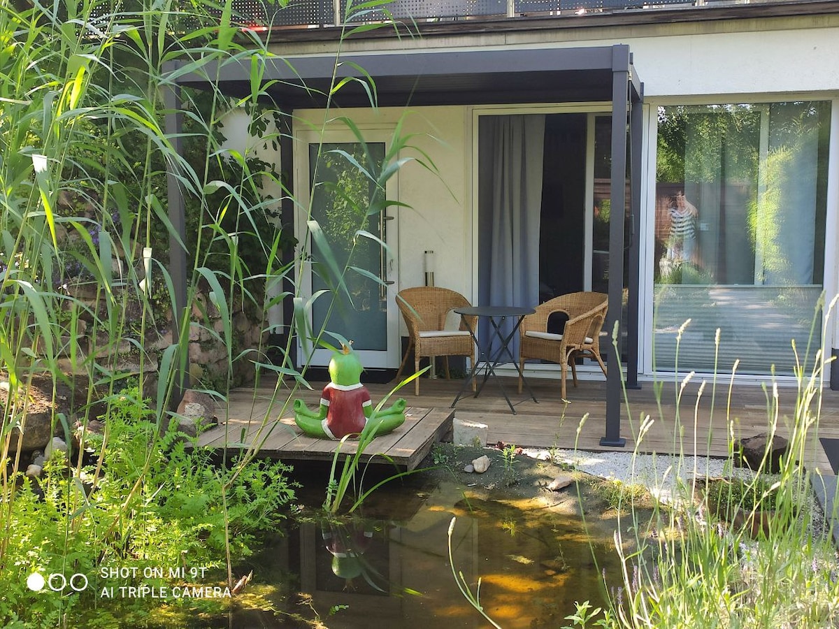 A private terrace is presented with two wicker chairs and a small table, situated next to a tranquil pond. Lush greenery surrounds the area, enhancing the serene environment. Large glass doors suggest a seamless connection to the indoor living space.