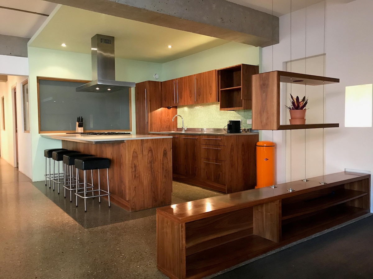 A modern kitchen features wooden cabinets with a light green backsplash and stainless steel appliances. A central island is accompanied by sleek bar stools. A vibrant orange appliance adds a pop of color to the space, while a minimalist wooden shelf displays a potted plant.
