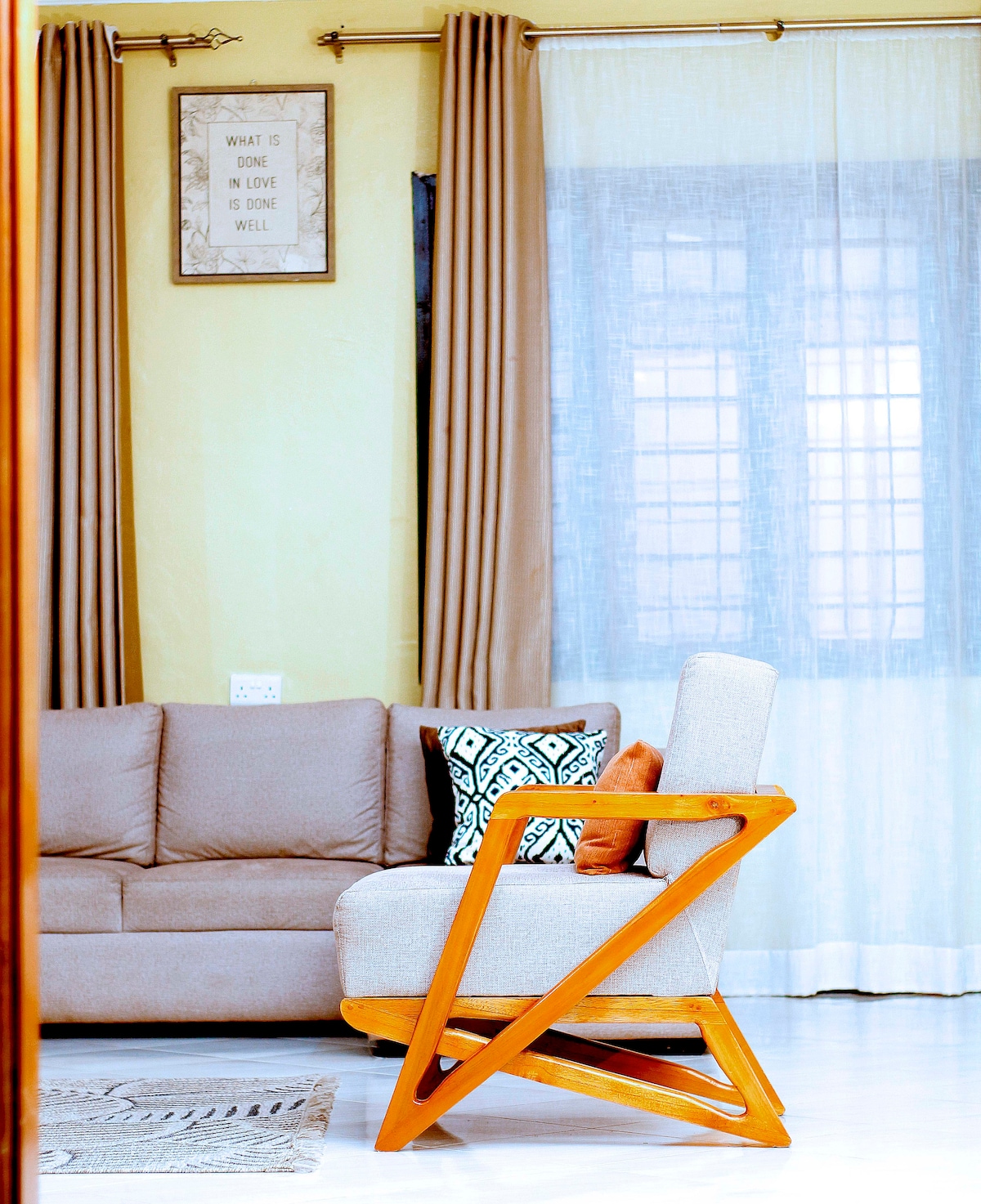 A comfortable living space features a light-colored sofa paired with decorative pillows. A stylish wooden chair with an angular design is positioned nearby. Sheer curtains allow natural light to filter in, softening the ambiance, while wall art adds a decorative touch.