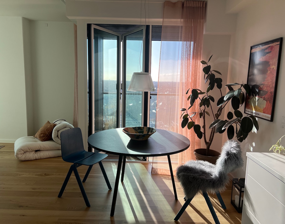 A dining area is depicted featuring a round table with a decorative bowl at its center. A blue chair and a furry grey chair are positioned on either side. Natural light streams in through large floor-to-ceiling windows, complementing a nearby potted plant and soft floor cushions.