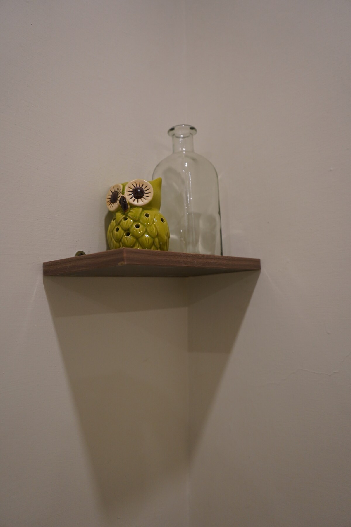 A small wooden shelf is mounted on a plain wall. A yellow ceramic owl and a clear glass bottle are positioned on the shelf, creating a simple yet charming display.
