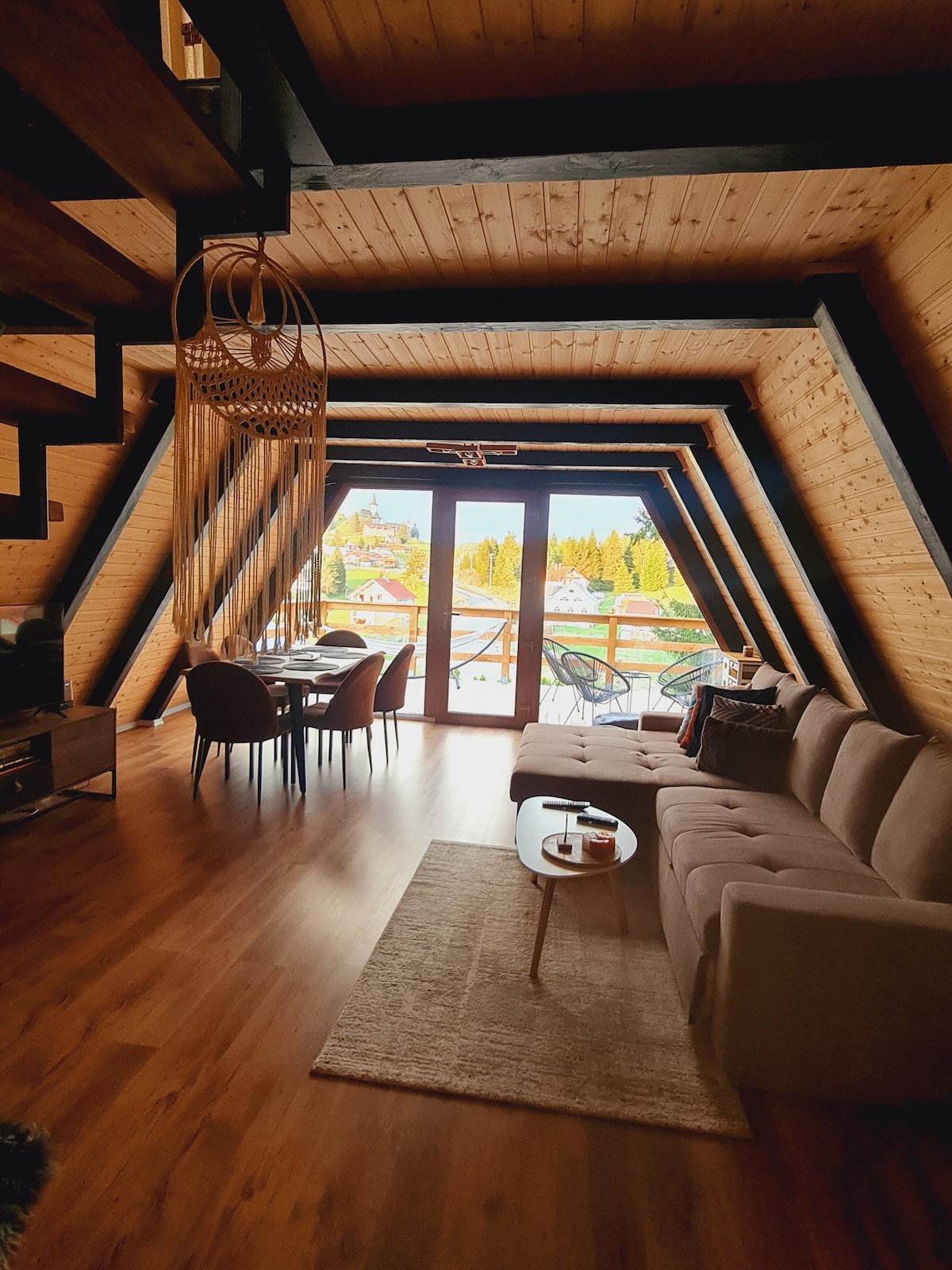 An open living space is revealed, featuring a modern couch and a round dining table surrounded by stylish chairs. Large glass doors frame a scenic view of the outdoors, while wooden paneling contrasts with the black beams overhead. A soft area rug adds comfort to the floor.