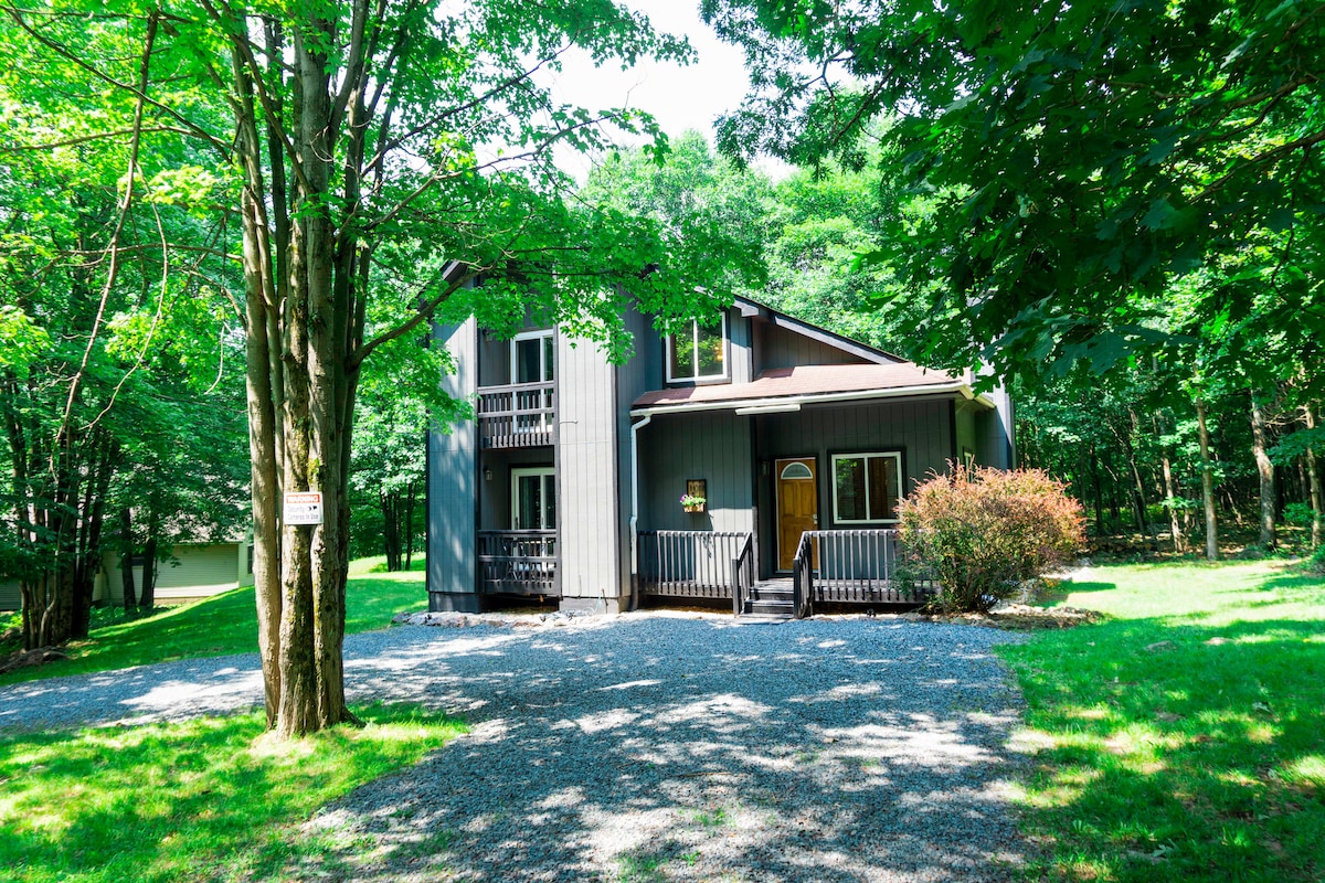The two-story cottage is surrounded by lush greenery, featuring a combination of wooden and gravel pathways. A spacious front porch with decorative railing is visible, inviting guests to enjoy nature. Large windows allow for ample natural light, enhancing the tranquil outdoor atmosphere.