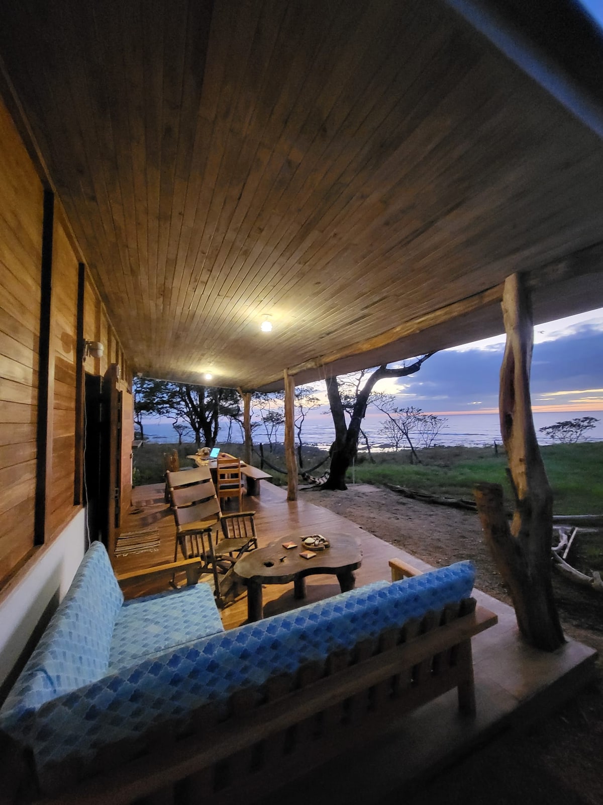 A spacious terrace features a comfortable seating area with a light blue sofa and wooden chairs. A wooden table is set for dining. Natural wood beams support the ceiling, and the landscape is visible in the background, with the sky transitioning into evening colors.