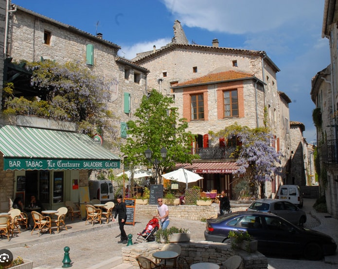 A quaint village square is marked by stone buildings featuring wood-trimmed windows and vibrant greenery. Outdoor seating is arranged at nearby cafes under umbrellas. Wisteria blooms add color to the scene, while people stroll along the cobblestone path. A parked car is visible in the foreground.
