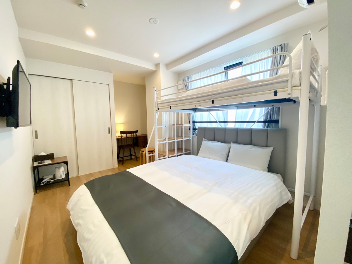 A contemporary room features a spacious double bed with white linens and a gray accent, complemented by a bunk bed above. Natural light filters through the sheer curtains, illuminating the wooden floor. A TV is mounted on the wall, and a small dining table with chairs is visible.
