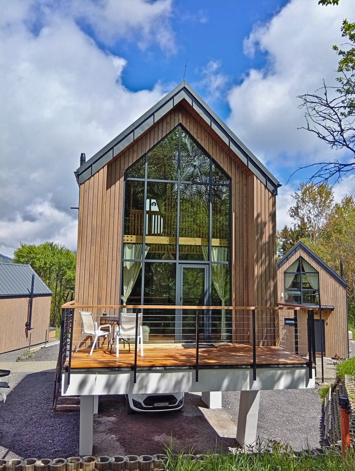 A modern cabin is elevated on pillars, featuring a spacious wooden deck with white outdoor furniture. Large glass panels dominate the front, allowing for ample natural light. Set against a backdrop of greenery and a partly cloudy sky, the structure blends contemporary design with nature.