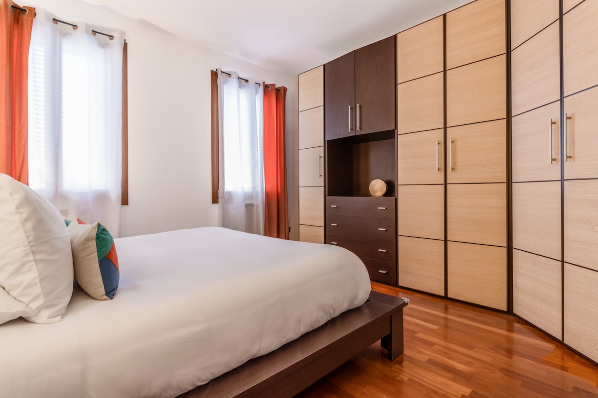 A spacious bedroom is highlighted by a large bed with a white duvet and decorative pillows. Natural light filters through multiple windows with light curtains. A modern wardrobe with a mix of wood finishes provides ample storage, enhancing the room's functionality.