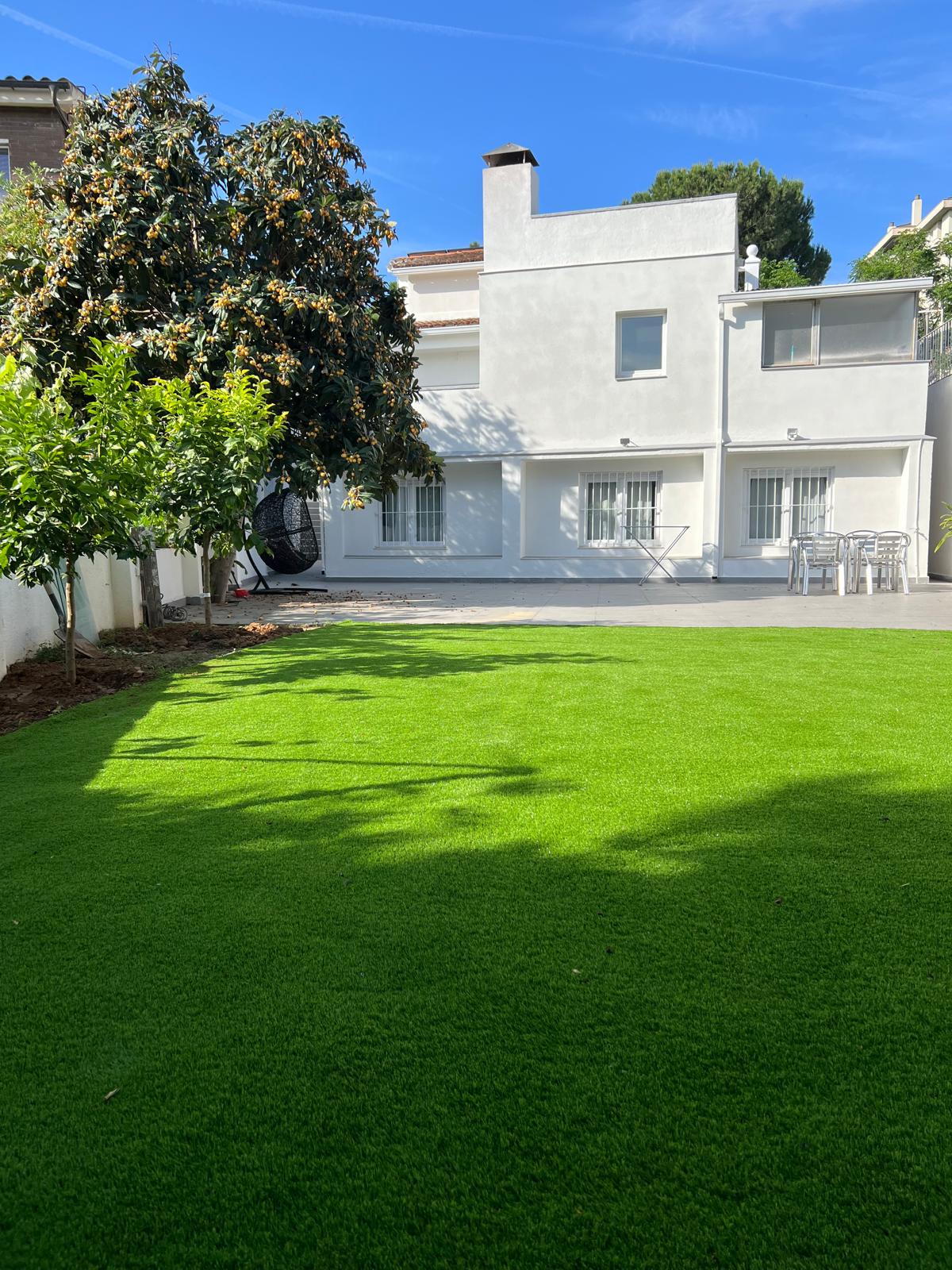 A private backyard area is shown, featuring lush green artificial turf that creates a neat and well-maintained appearance. A white two-story building with multiple windows forms the backdrop, while a dining set with multiple chairs is visible in the space.