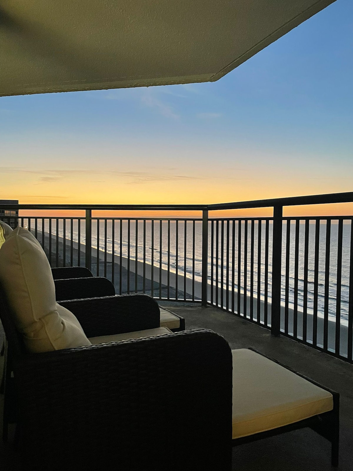 A spacious balcony offers stunning ocean views at sunrise, enhanced by comfortable patio chairs. The railing provides a clear view of the shoreline, creating an inviting space for relaxation while overlooking the beach.