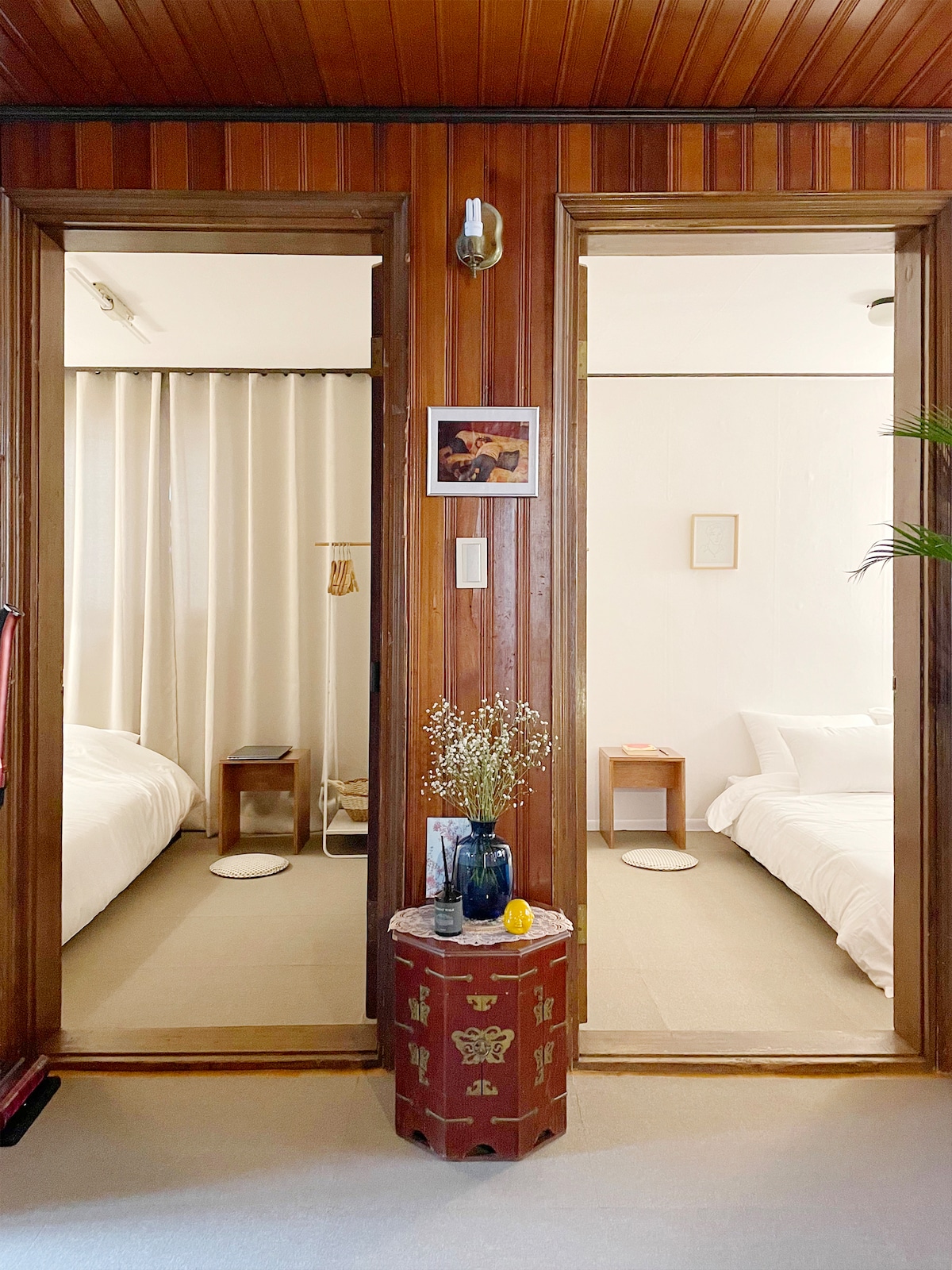 A wooden entryway connects two bedrooms, each with a distinct aesthetic. Soft curtains and minimal decorations enhance the calming atmosphere. A small side table adorned with flowers and a decorative vase stands in the foreground, adding a touch of charm to the space.