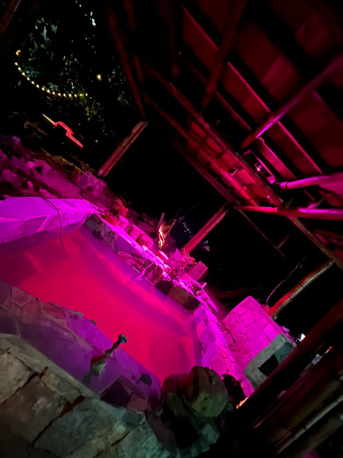 A stone jacuzzi with warm, flowing water is surrounded by natural rock features. Soft pink lighting creates a calming ambiance, enhancing the tranquil outdoor setting. The jacuzzi is covered by a rustic structure, providing a cozy atmosphere for relaxation.