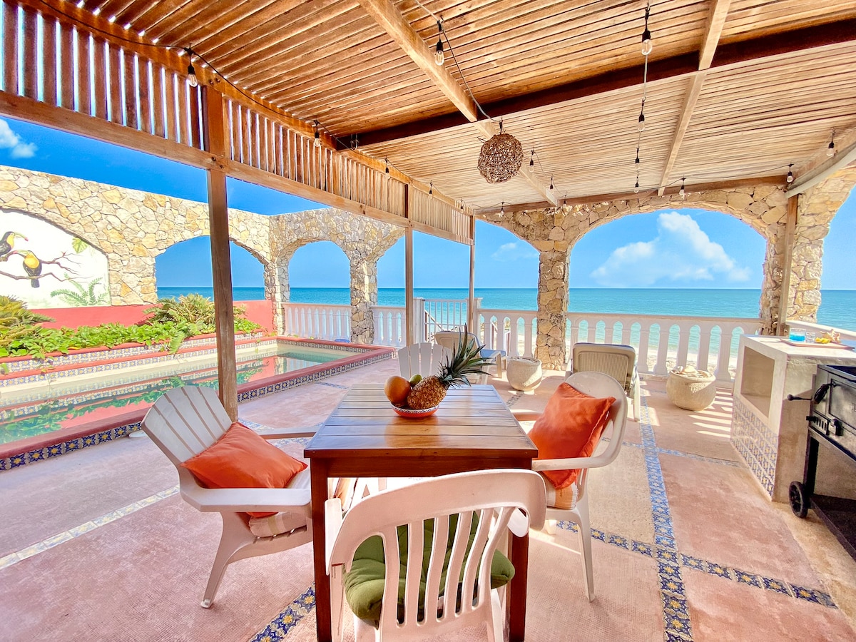 An outdoor dining area is situated under a wooden pergola, featuring a wooden table surrounded by comfortable chairs. Stone arches frame the view of the ocean, with a colorful garden in front. Bright sunlight highlights the tranquil setting by the water.