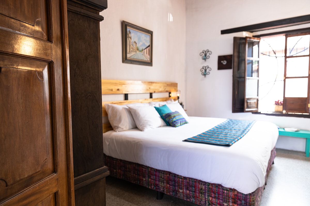 A spacious bedroom features a plush queen bed adorned with a colorful blanket. Natural light filters through the window, highlighting the wooden furniture and a painting on the wall. A traditional ambiance is created by the blend of textures and warm tones.