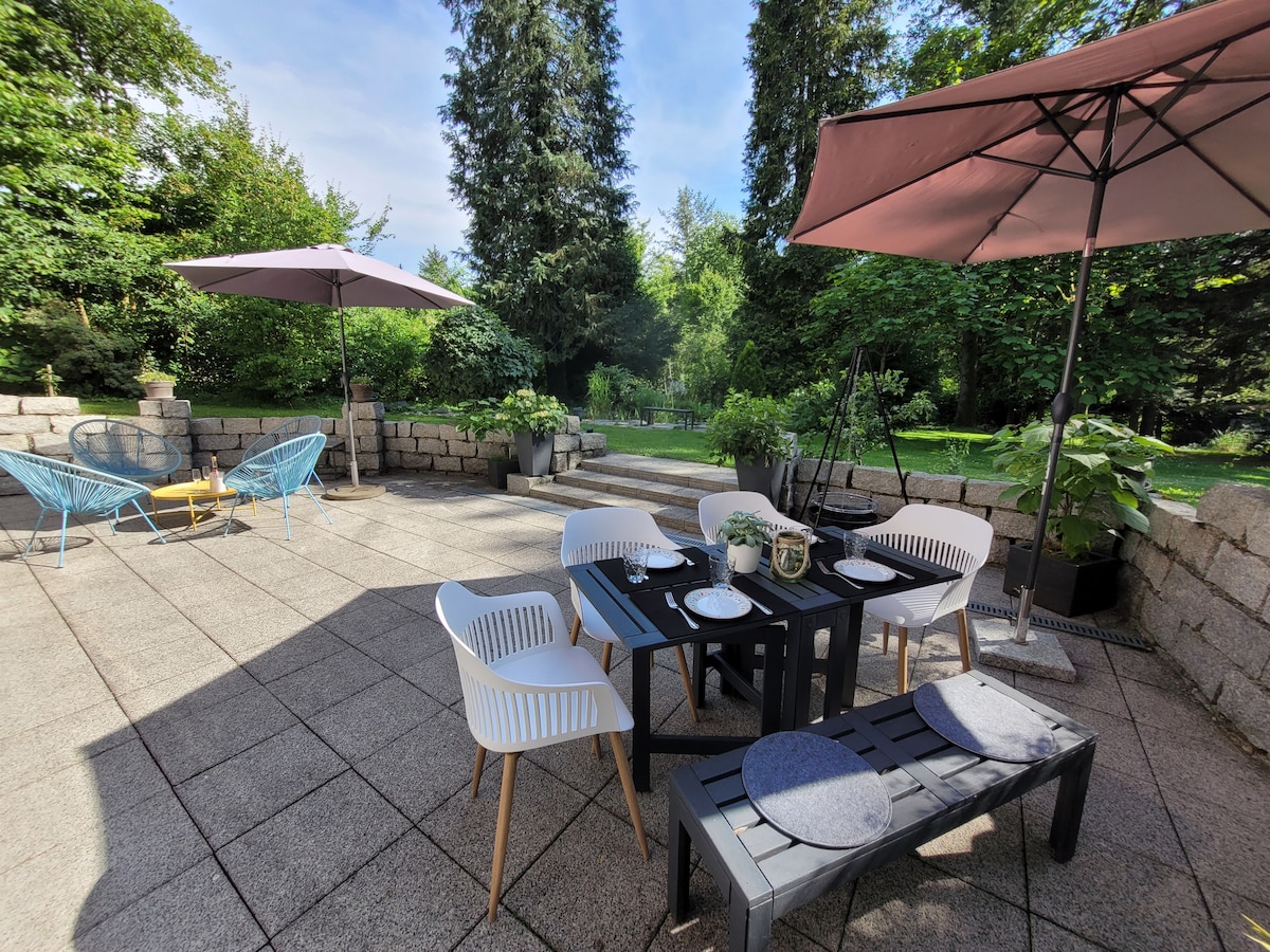 A generous terrace is featured with a dining table surrounded by six chairs. Two umbrellas provide shade, while a seating area includes lounge chairs and a swing. The space opens onto a lush garden, enhancing outdoor relaxation with a tranquil atmosphere.
