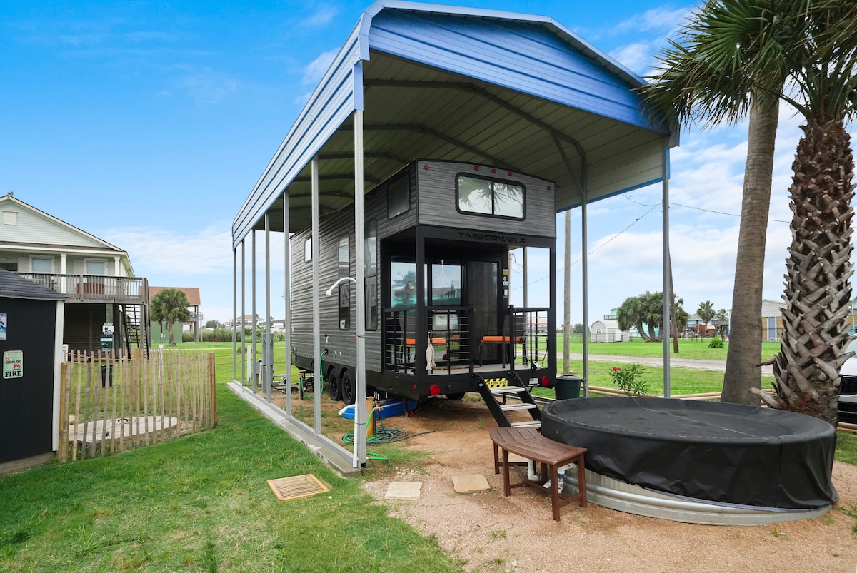 The image captures a tiny home situated under a blue roofed canopy, with a covered outdoor area visible. A private plunge pool is situated to the side, surrounded by neatly trimmed grass. Nearby palm trees add to the relaxing outdoor atmosphere.