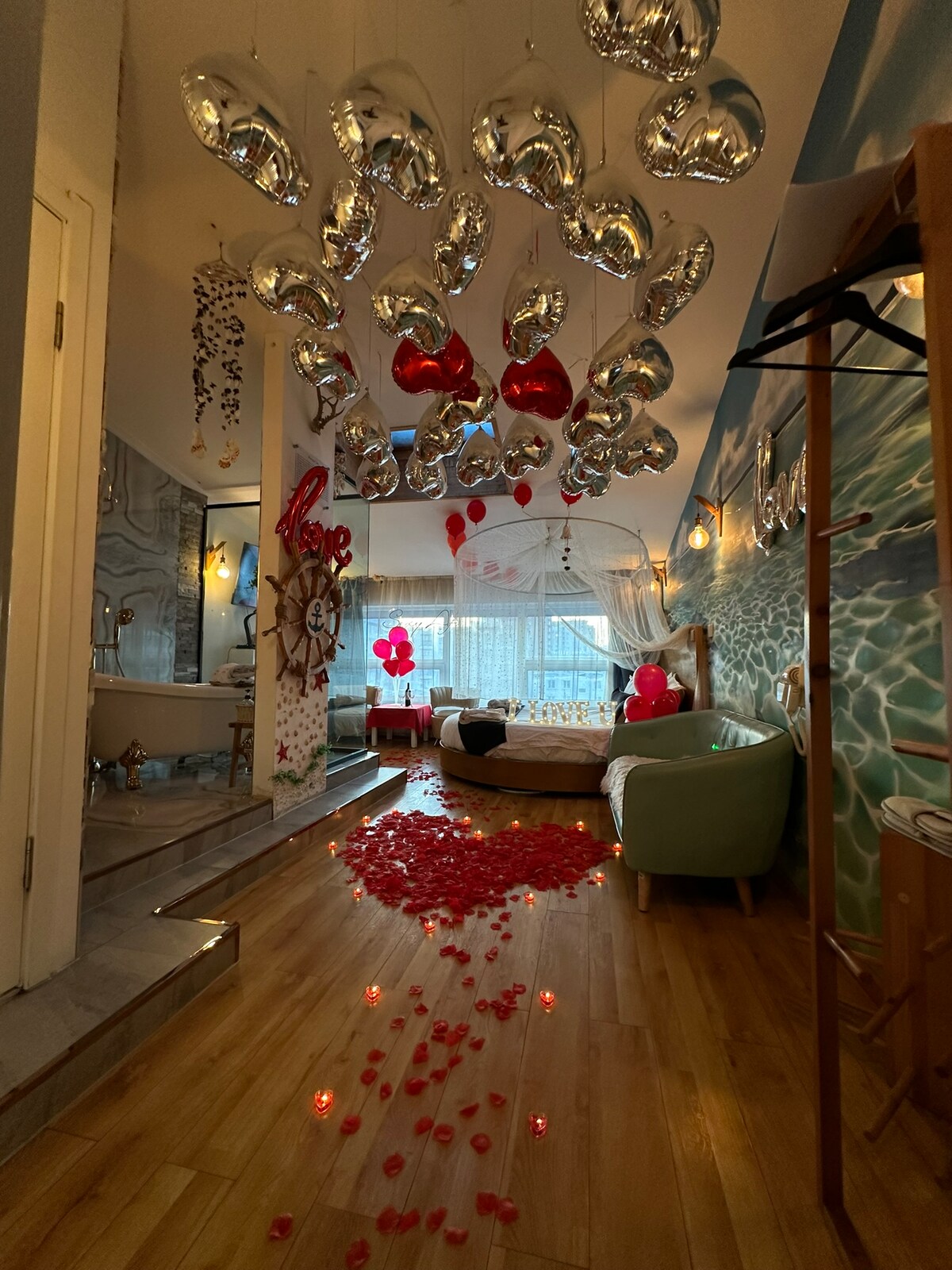 A whimsical interior showcases a variety of heart-shaped balloons suspended from the ceiling. A pathway of rose petals leads to a cozy seating area with a light blue armchair. Natural light filters through sheer curtains, illuminating the ocean-themed wall mural.