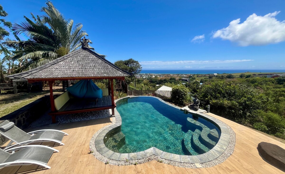 A serene outdoor area features a curved swimming pool with clear water reflecting the sky. A traditional gazebo is positioned nearby, along with several lounge chairs. Lush greenery surrounds the space, providing a calming environment, complemented by a distant view of the sea.