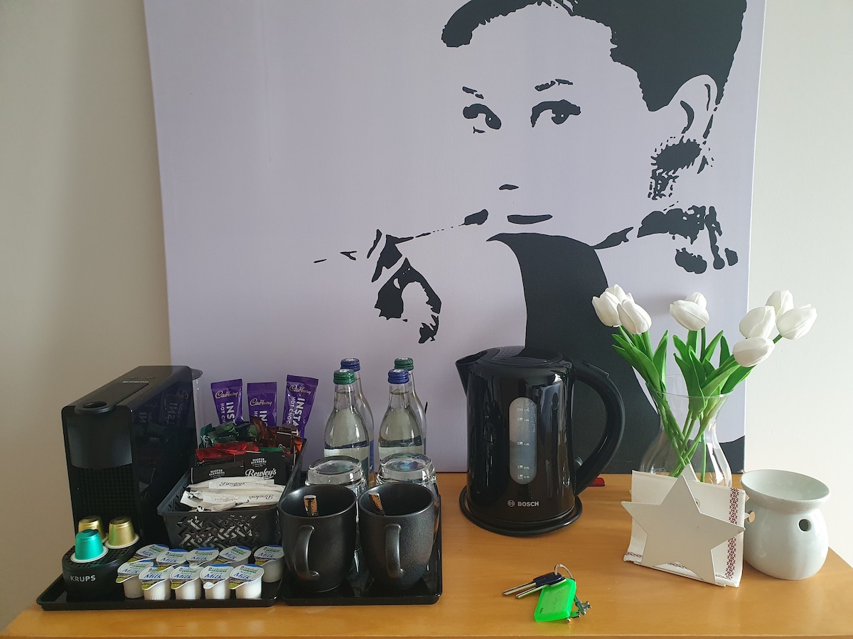 A coffee station is set up with an espresso machine, a kettle, and two mugs. Bottled water is paired with coffee and tea supplies, including pods and sugar packets. White tulips and a decorative vase add a touch of freshness to the arrangement against a wall featuring a black and white design.