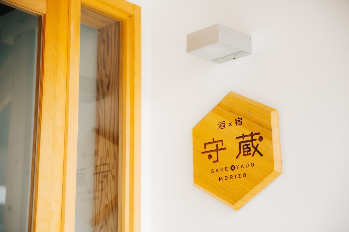 A wooden sign featuring the name '守蔵' is displayed on a white wall, illuminated by a light fixture above. The sign has a hexagonal shape and combines classic typography with a modern design, emphasizing the connection between sake and lodging.