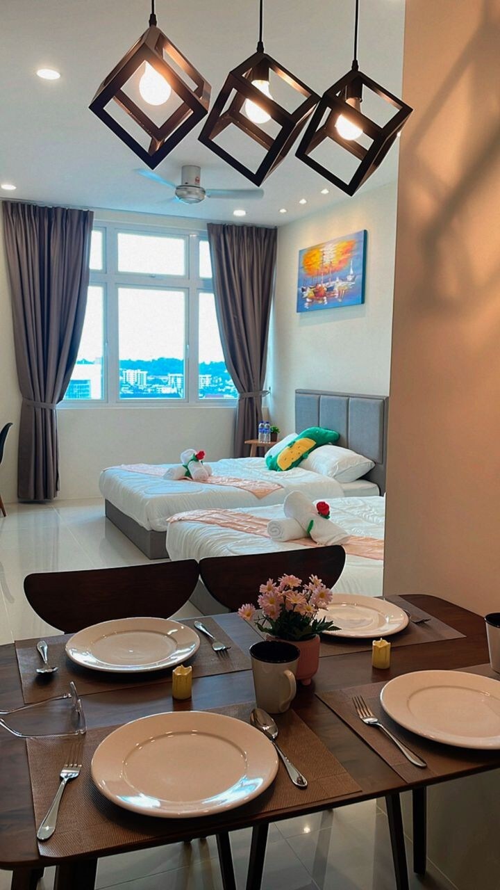 A studio unit is displayed, featuring two spacious beds adorned with decorative pillows and a vibrant painting above. A dining table set for four is in the foreground, complemented by natural light streaming in through large windows with curtains.