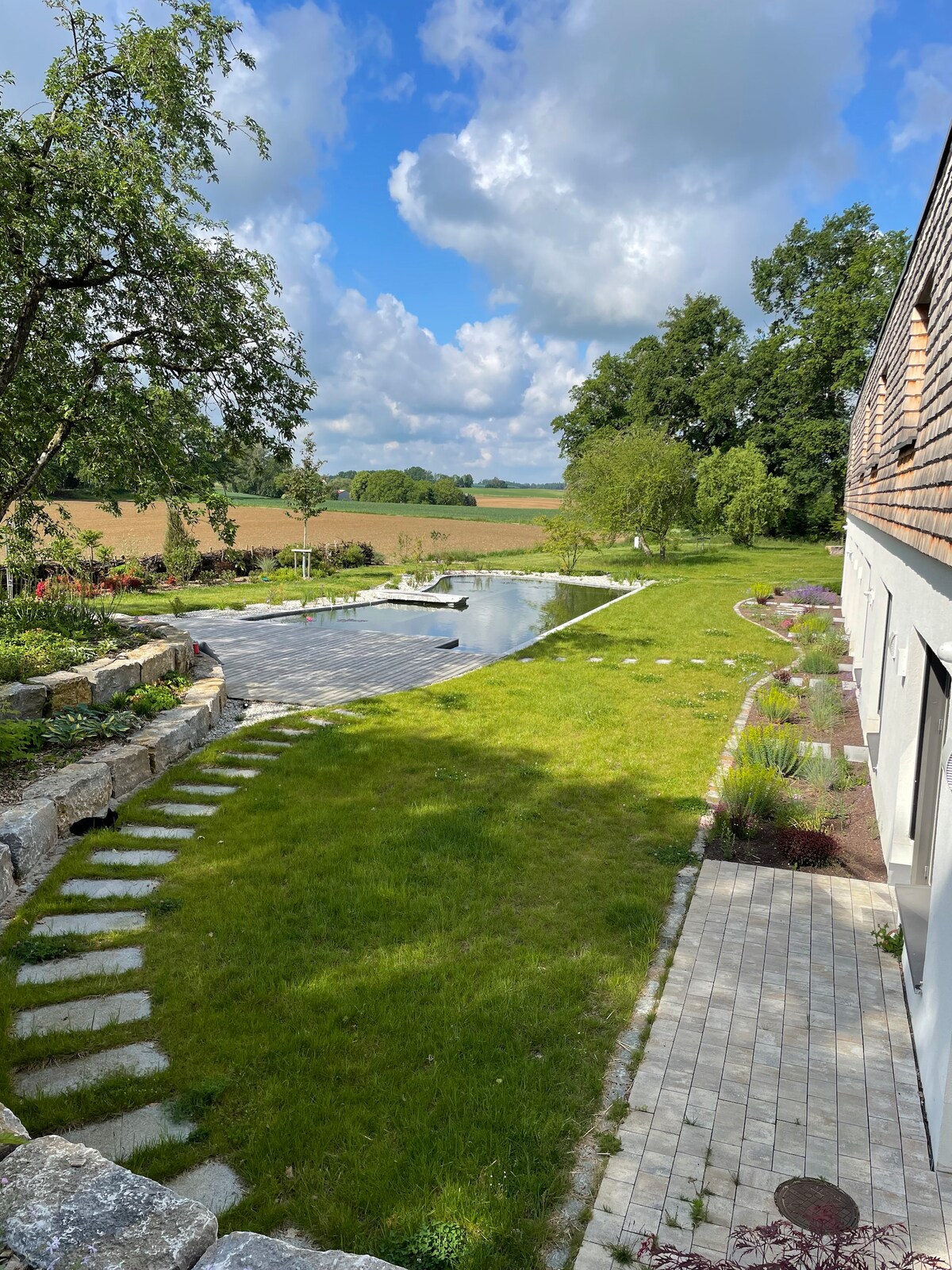 A serene outdoor area features a natural pool surrounded by lush green grass and gardens. Stone pathways lead through the space, inviting exploration. Cloudy skies above reflect a peaceful atmosphere, while fields stretch into the distance, providing a sense of openness and tranquility.