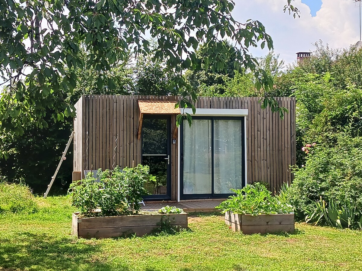 A modern wooden studio is nestled in a garden, surrounded by lush greenery. Large glass doors provide natural light and connect the interior to the outdoor space. Raised garden beds with plants are visible in the foreground, blending with the tranquil environment.