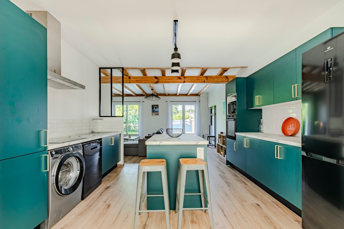 A modern kitchen is presented with teal cabinetry and sleek white countertops. Stainless steel appliances are visible, including a washer and dryer. An island with two stools is positioned centrally, while large windows allow natural light to flood the space, creating an airy atmosphere.