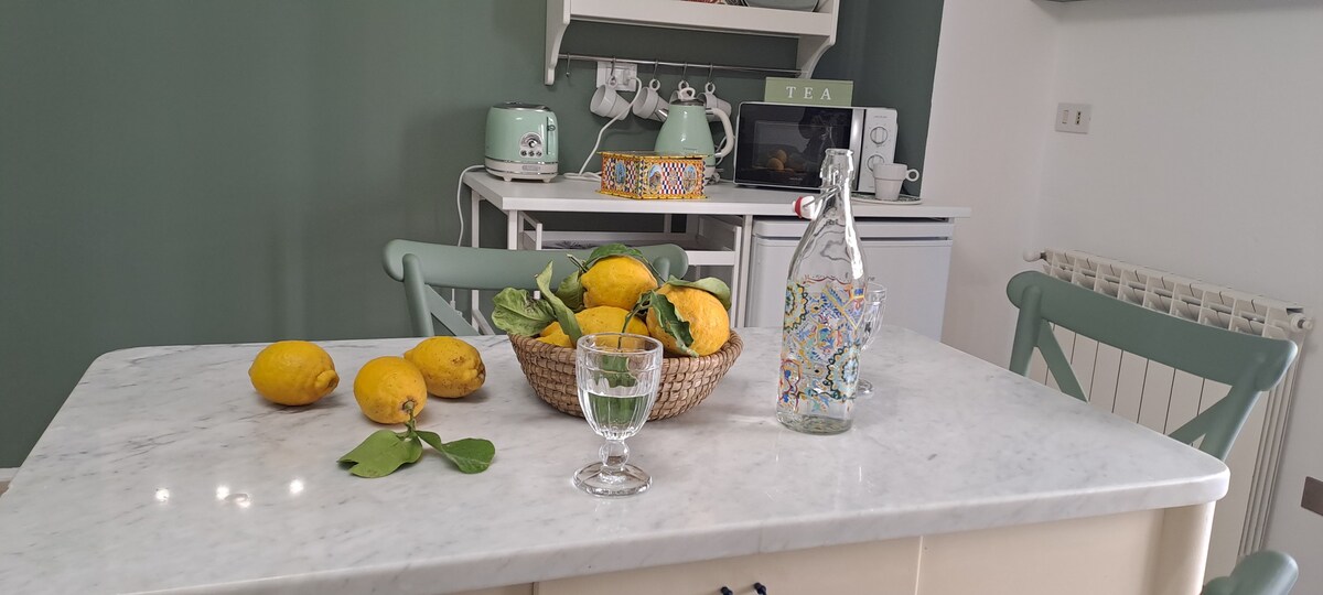 A light marble table holds a woven bowl filled with fresh lemons, accompanied by a glass of water. A decorative glass bottle sits nearby, adding to the serene kitchen setting, which features green walls and modern kitchen appliances in the background.