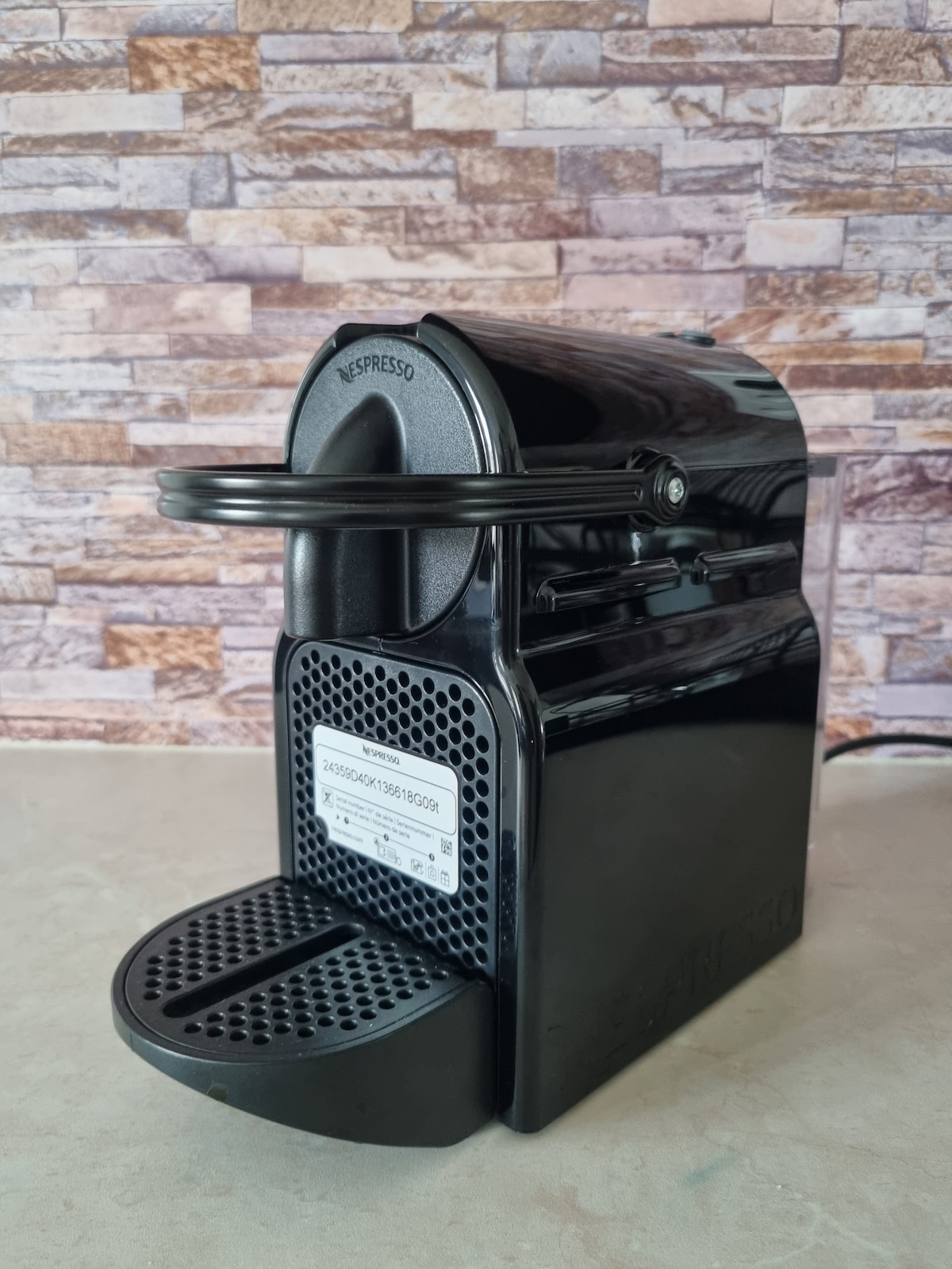 A sleek black coffee machine is positioned on a light-colored surface, featuring a textured drip tray and a simple operation lever. The backdrop consists of a stone wall with varying shades, adding a modern touch to the kitchen space.