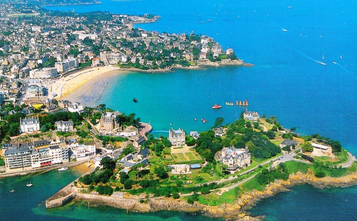 An aerial view captures the coastal beauty of Dinard, showcasing sandy beaches and vibrant blue waters. The image reveals a mix of urban and natural landscapes, with distinct buildings lining the shore and greenery surrounding the area, providing a sense of tranquility by the sea.