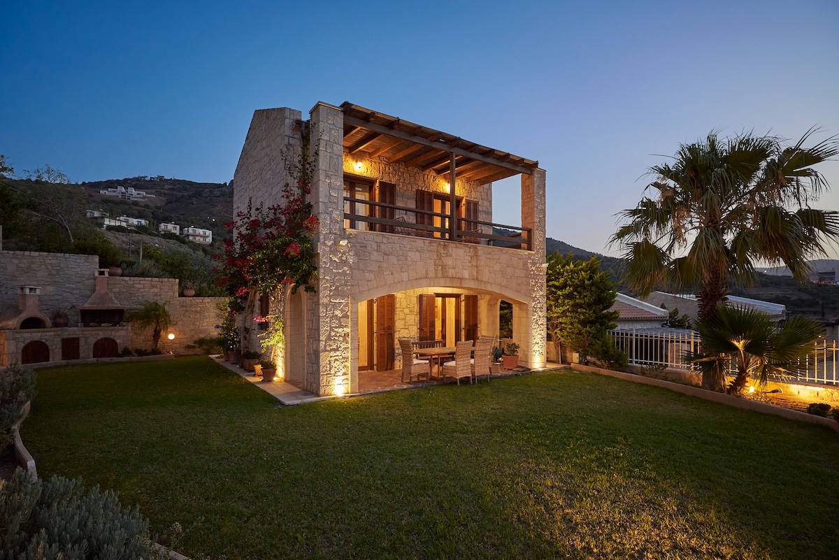 A stone villa is set in a garden, featuring a spacious outdoor area illuminated softly in the evening light. The upper balcony showcases wooden accents, while vibrant greenery and flowering plants surround the structure, creating a serene and inviting outdoor space.