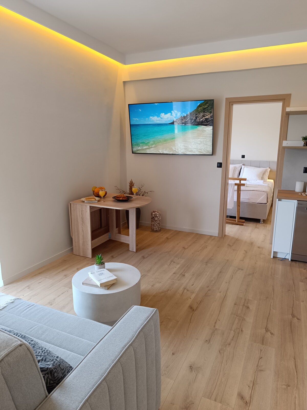 A light-filled living area features a comfortable sofa and a small white table. A mounted screen displays a serene beach scene, enhancing the relaxing environment. A wooden console holds decorative items and snacks, while a doorway leads to the adjacent bedroom.