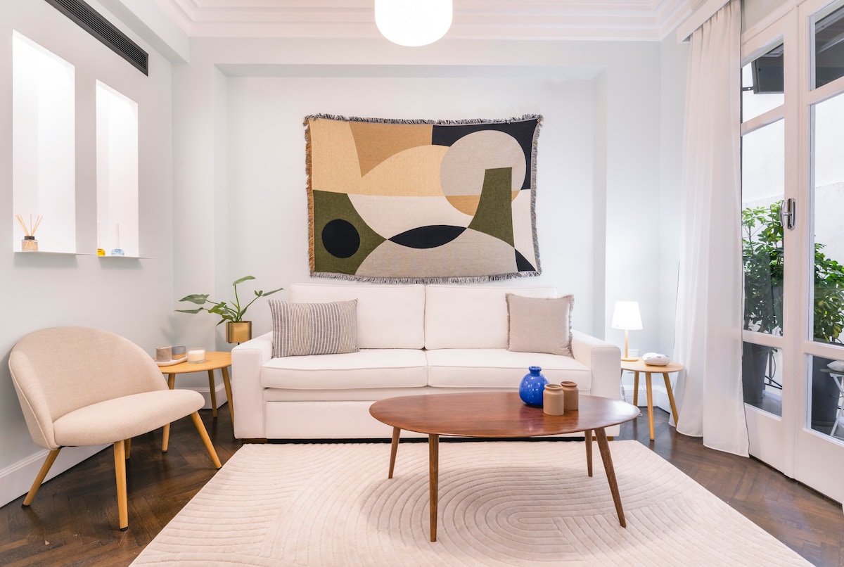 The living area features a light-coloured sofa with decorative cushions, complemented by a round wooden coffee table. A wall tapestry adds visual interest, while greenery is visible through large glass doors leading to a balcony. Soft lighting creates a cozy ambiance.