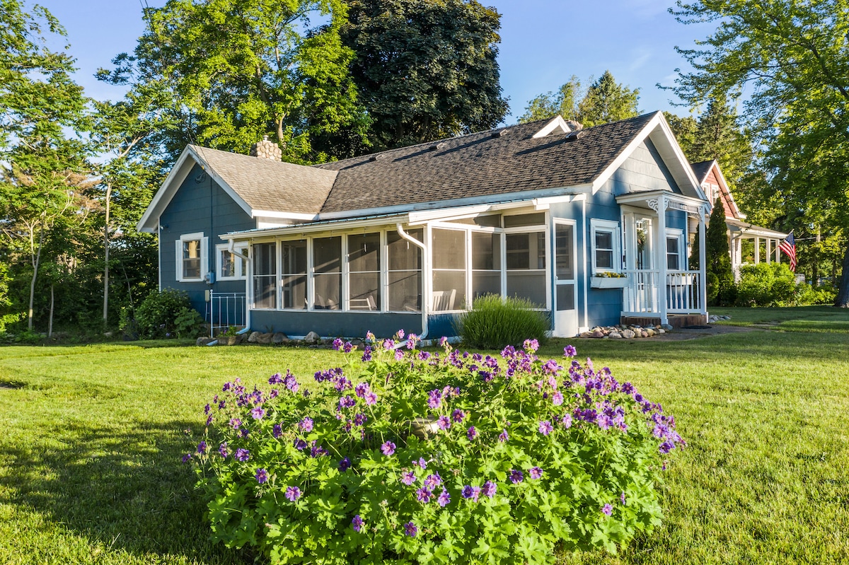 A blue cottage is surrounded by lush green grass, featuring a screened porch that offers outdoor views. Vibrant purple flowers are planted in the foreground, complementing the serene garden setting. Tall trees provide a natural backdrop, enhancing the peaceful environment of the property.