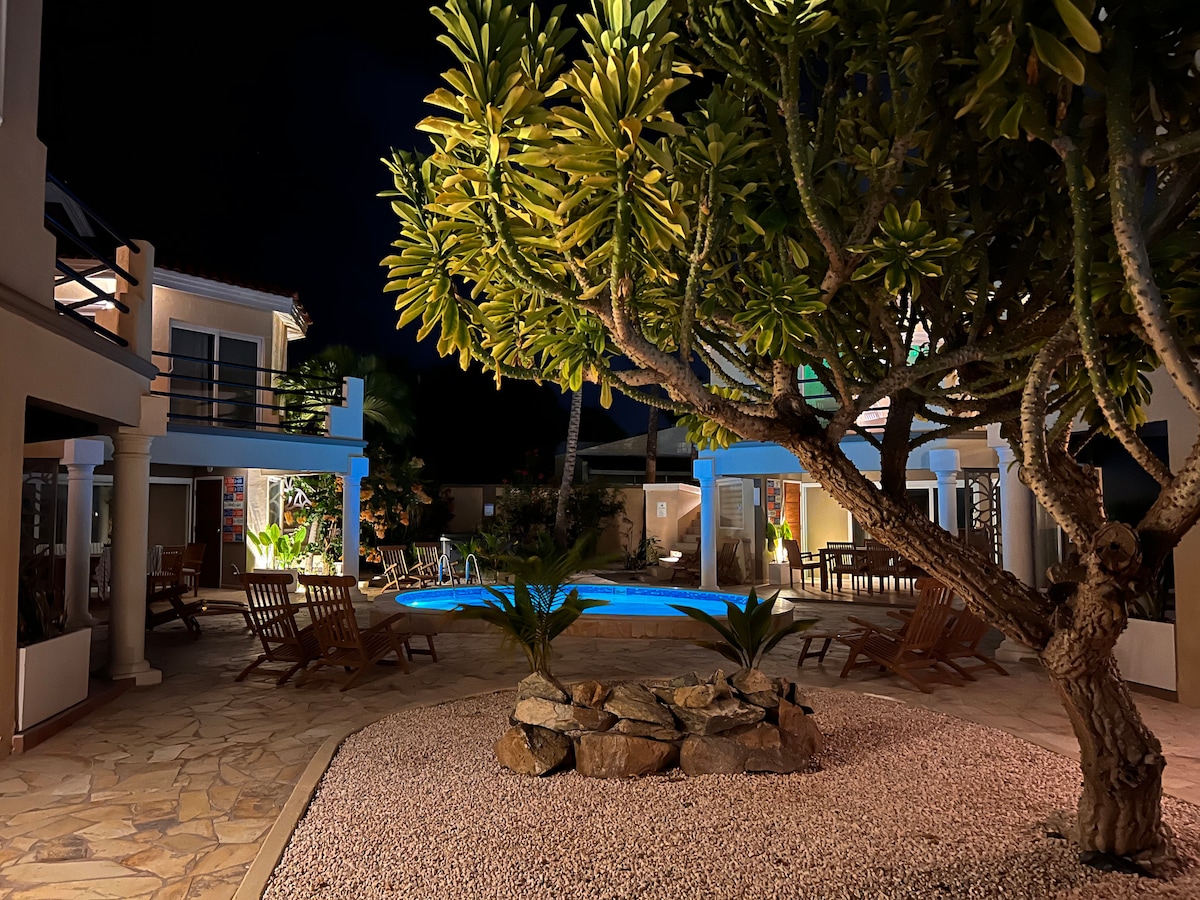 The pool area is illuminated at night, surrounded by lush greenery and stone landscaping. Comfortable wooden lounge chairs are positioned around the pool, creating inviting spaces for relaxation. The villas are softly lit, enhancing the tranquil atmosphere of the outdoor retreat.