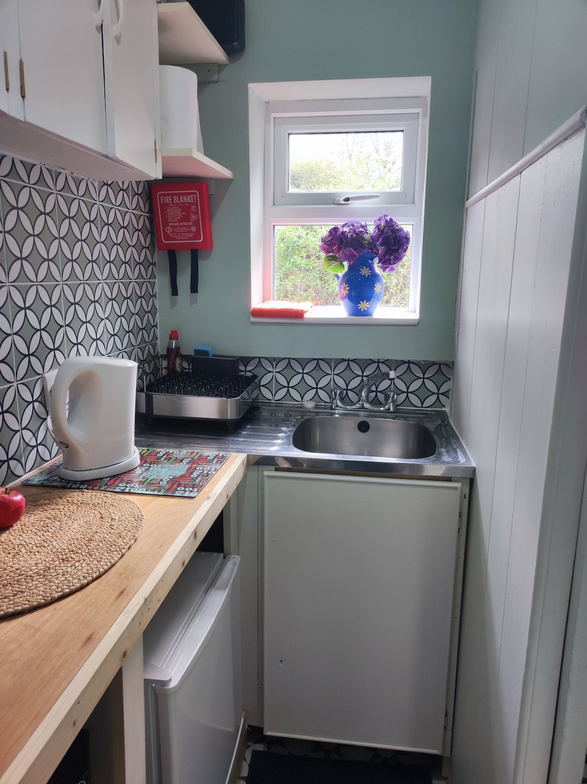 The compact kitchen features a stainless steel sink and a small refrigerator, complemented by a kettle and dish rack. A window allows natural light to filter in, framed by a vase of purple flowers. Decorative tiles add a touch of pattern to the wall.