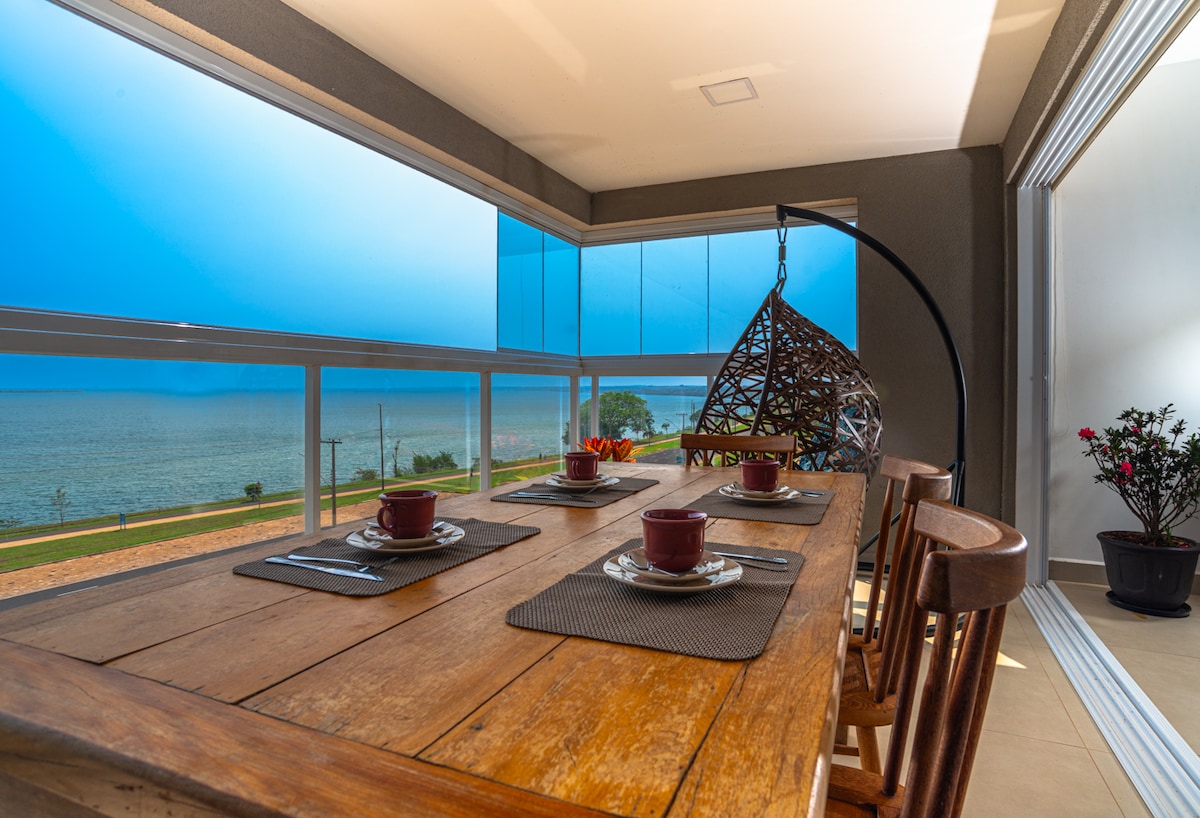 A dining table made of wood is set with cups on placemats, positioned in a well-lit area. A hanging chair adds an inviting touch. Large windows offer expansive views of the water and greenery, allowing natural light to fill the space.