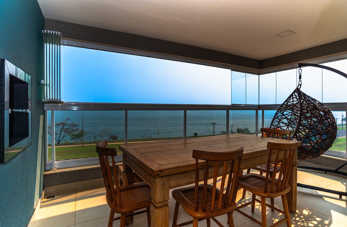 A dining area features a wooden table surrounded by sturdy chairs, positioned next to large glass windows showcasing a panoramic view of the ocean. A hanging chair adds comfort, while the walls are painted in a calming blue tone.